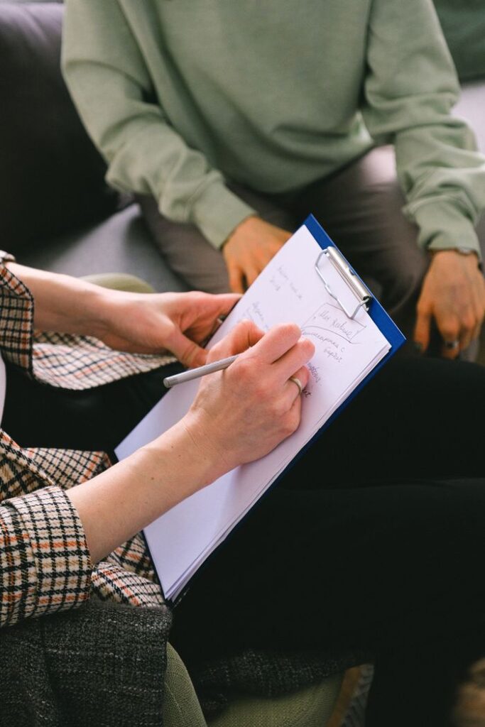 Home Crop anonymous female coach writing notes on paper in clipboard while talking with client in room